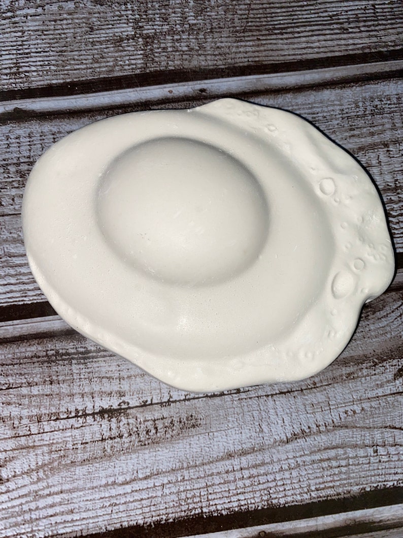 May include: A white ceramic dish shaped like a fried egg. The dish has a rounded center and a wavy edge with small, circular indentations. The egg-shaped dish is on a wooden surface.