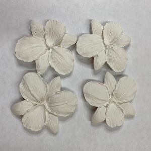 May include: Four off-white, textured flower-shaped decorative pieces. Each flower has multiple petals and a detailed center. The pieces are arranged in a square formation against a neutral background.