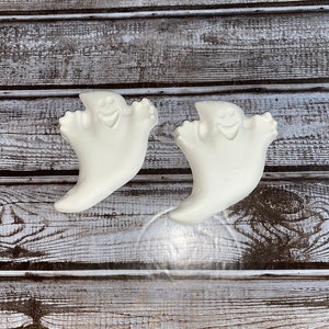 May include: Two white ghost-shaped decorations with smiling faces and outstretched arms. The ghosts are made of a smooth material and are set against a weathered, gray and white wooden background. These are likely Halloween decorations.