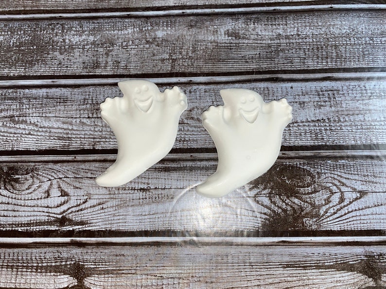 May include: Two white ghost-shaped decorations with smiling faces and outstretched arms. The ghosts are made of a smooth material and are set against a weathered, gray and white wooden background. These are likely Halloween decorations.