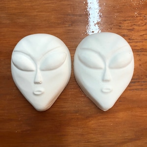 May include: Two white ceramic alien face sculptures with closed eyes and a small mouth. The sculptures are on a wooden surface.