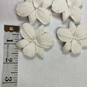 May include: Four off-white, flower-shaped decorative pieces. Each flower has five petals and a textured surface. A measuring tape shows the flowers are approximately 2 inches in diameter.