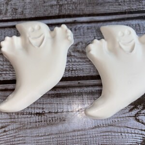 May include: Two white ghost-shaped decorations with smiling faces. The ghosts have a rounded shape with outstretched arms. The background is a weathered, gray wooden surface. These are Halloween-themed decorations.