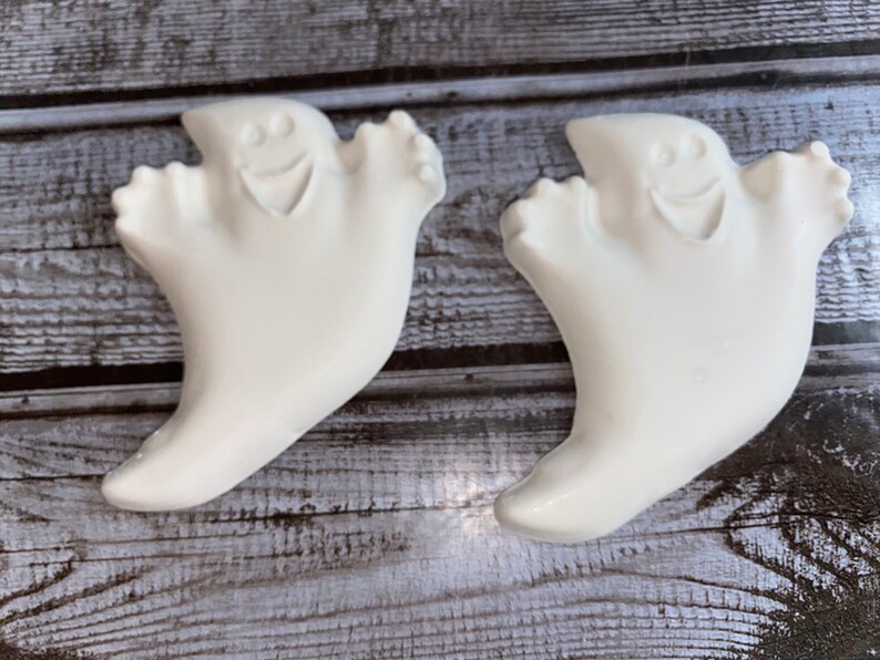 May include: Two white ghost-shaped decorations with smiling faces. The ghosts have a rounded shape with outstretched arms. The background is a weathered, gray wooden surface. These are Halloween-themed decorations.