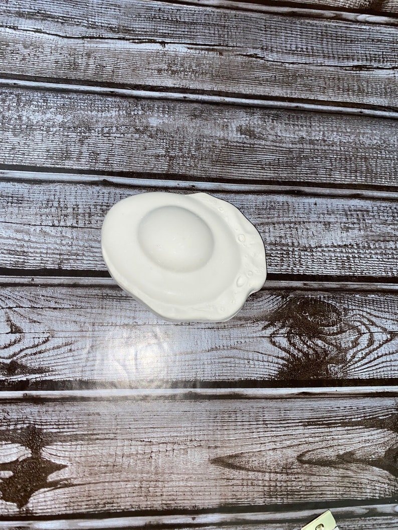 May include: A white ceramic egg-shaped dish with a slightly irregular edge. The dish has a smooth, glossy surface and a shallow, rounded center. The design mimics a fried egg, perfect for serving small portions or as a decorative accent.