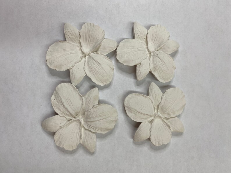 May include: Four off-white, sculpted flower ornaments. Each flower has five petals and a detailed center. The flowers are arranged in a square pattern against a neutral background. Ideal for craft projects.