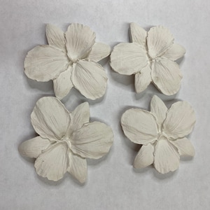 May include: Four off-white, sculpted flower ornaments arranged in a square. Each flower has detailed petals and a textured surface. The flowers are likely for decorative purposes, possibly for crafts or home decor.