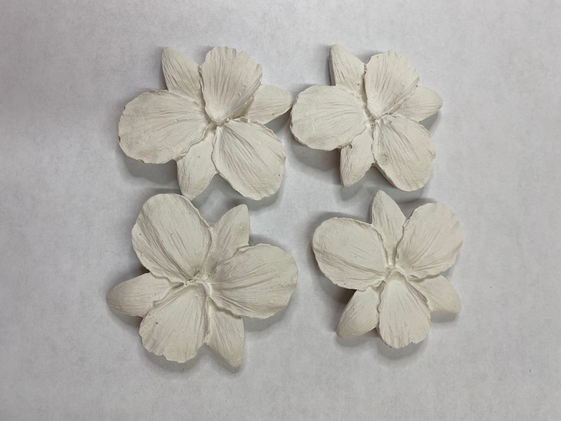 May include: Four off-white, sculpted flower ornaments arranged in a square. Each flower has detailed petals and a textured surface. The flowers are likely for decorative purposes, possibly for crafts or home decor.