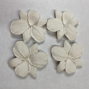 May include: Four off-white, textured flower-shaped decorative pieces. Each flower has multiple petals and a detailed center. The flowers are arranged in a square pattern against a neutral background. These could be used for craft projects or home decor.
