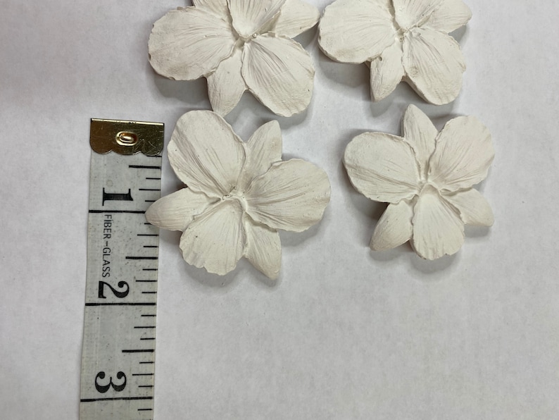May include: Four off-white, flower-shaped decorative pieces. Each flower has five petals and a textured surface. A measuring tape shows the flowers are approximately 2 inches in diameter.