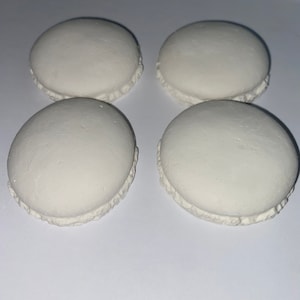 May include: Four white macarons on a white surface. The macarons are round and have a slightly ruffled edge.