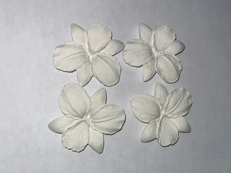 May include: Four white, flower-shaped decorative elements. Each flower has multiple petals and a detailed, textured surface. The objects are arranged on a white background, showcasing their delicate design and potential for craft projects.