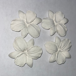 May include: Four white, flower-shaped decorative elements. Each flower has multiple petals and a detailed, textured surface. The objects are arranged on a white background, suggesting a craft or DIY project.