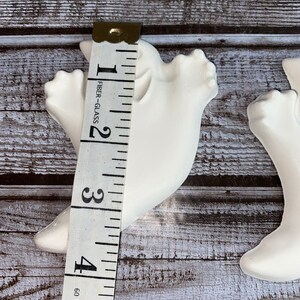 May include: Two white ceramic dove figurines, each approximately 4 inches tall, are shown with a measuring tape. The doves are positioned on a weathered, gray-toned wooden surface. The tape measure indicates the height of the dove.