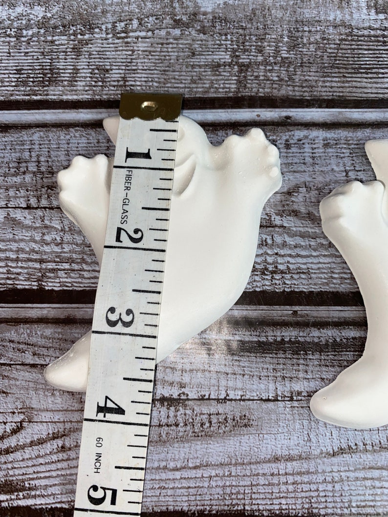 May include: Two white ceramic dove figurines, each approximately 4 inches tall, are shown with a measuring tape. The doves are positioned on a weathered, gray-toned wooden surface. The tape measure indicates the height of the dove.