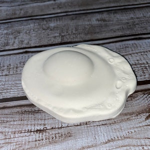 May include: A white, ceramic fried egg-shaped soap dish. The dish has a raised center resembling the yolk and a textured edge. The soap dish is designed to hold a bar of soap.