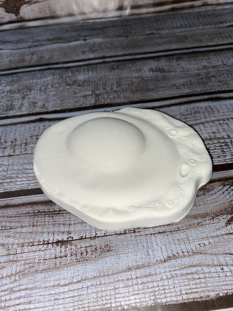 May include: A white, ceramic fried egg-shaped soap dish. The dish has a raised center resembling the yolk and a textured edge. The soap dish is designed to hold a bar of soap.