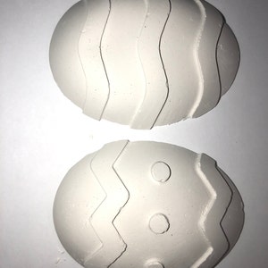 May include: Two white, egg-shaped decorations with textured designs. One has wavy lines, the other a zig-zag pattern with three small circles. These Easter-themed decorations are perfect for spring crafts and home decor.