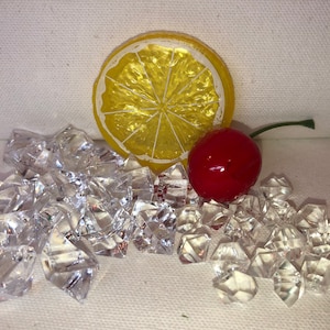 May include: A yellow lemon slice, a red cherry, and a pile of clear plastic ice cubes on a white background.