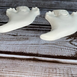 May include: Two white ceramic ghost figurines with a smiling face and outstretched arms. The ghosts are on a wooden surface with a weathered appearance. The figurines are likely for Halloween or seasonal decor.