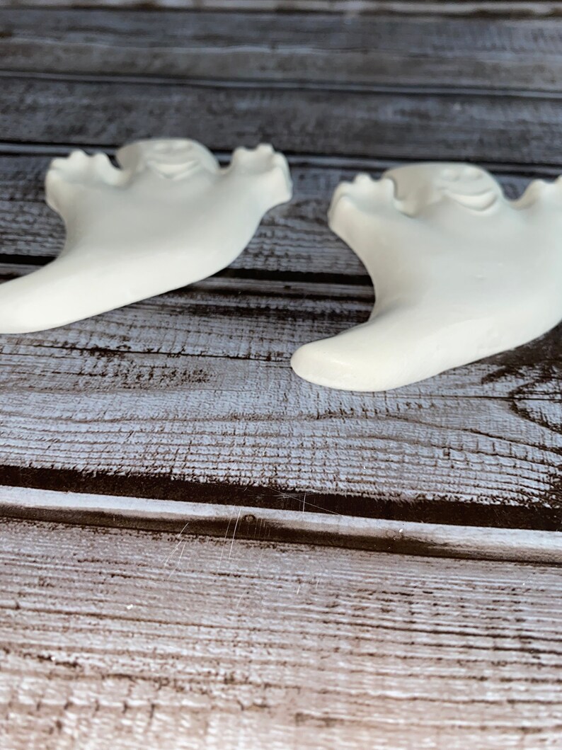 May include: Two white ceramic ghost figurines with a smiling face and outstretched arms. The ghosts are on a wooden surface with a weathered appearance. The figurines are likely for Halloween or seasonal decor.