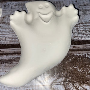 May include: A white ghost-shaped decoration with a smiling face and outstretched arms. The ghost has a smooth, rounded form and is likely made of ceramic or a similar material. The background is a wooden surface.