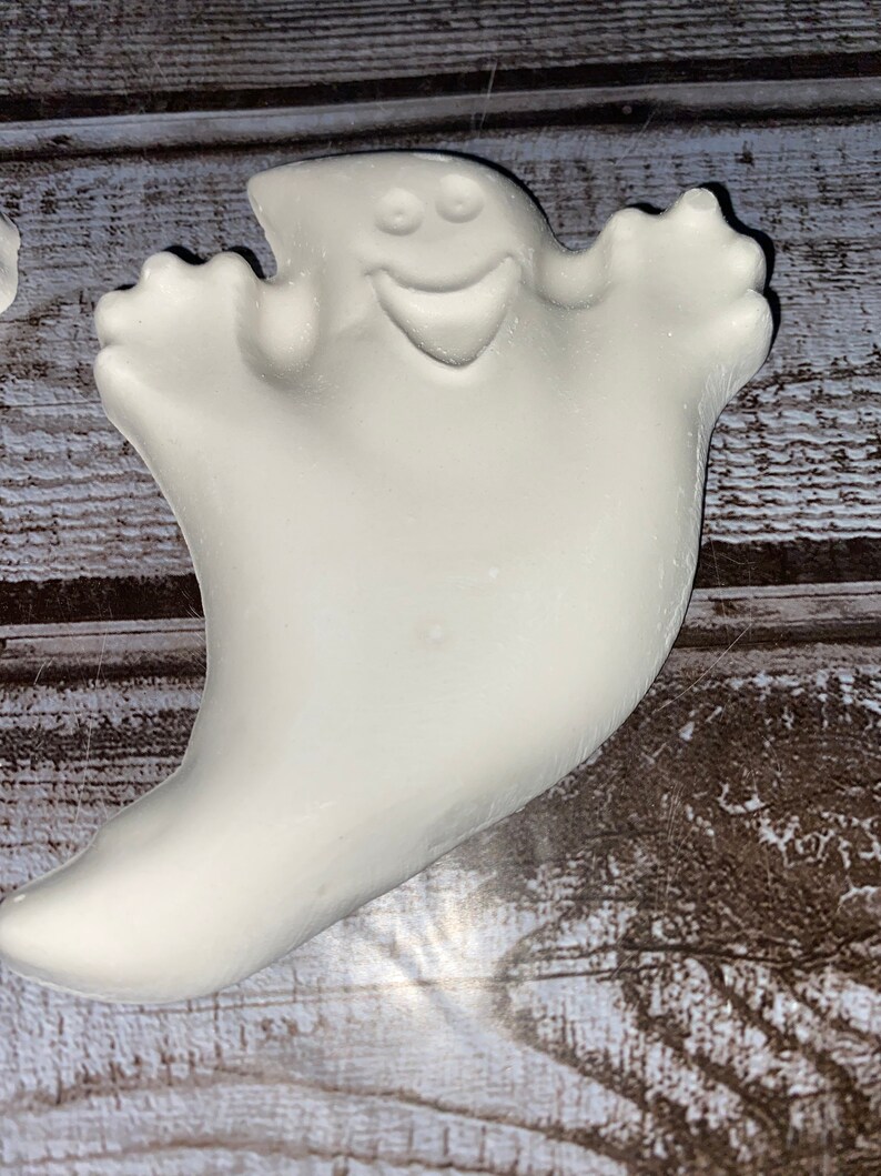 May include: A white ghost-shaped decoration with a smiling face and outstretched arms. The ghost has a smooth, rounded form and is likely made of ceramic or a similar material. The background is a wooden surface.