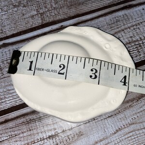 May include: A white ceramic dish with a circular indentation and a textured, irregular rim. The dish is approximately 4 inches in diameter. The dish is on a wooden surface.