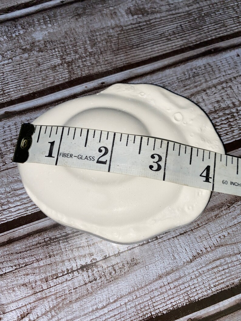 May include: A white ceramic dish with a circular indentation and a textured, irregular rim. The dish is approximately 4 inches in diameter. The dish is on a wooden surface.