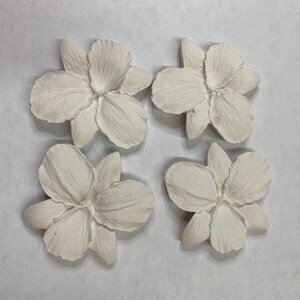 May include: Four off-white, textured flower-shaped decorative pieces. Each flower has multiple petals and a detailed center. The pieces are arranged in a square formation against a neutral background.