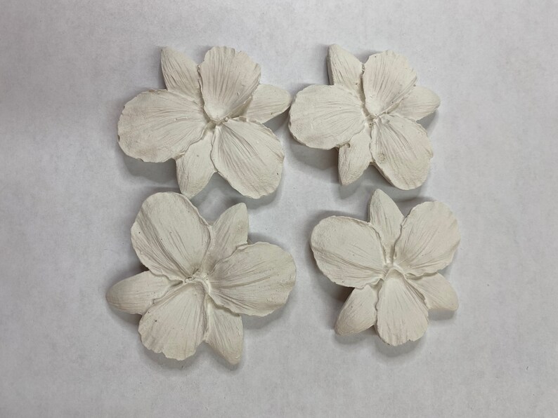 May include: Four off-white, textured flower-shaped decorative pieces. Each flower has multiple petals and a detailed center. The pieces are arranged in a square pattern against a light background. These could be used for craft projects or home decor.