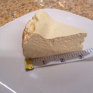 May include: A slice of cheesecake on a white plate, with a measuring tape showing a width of approximately 3 inches. The cheesecake has a light beige color and a textured surface.