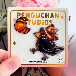 May include: A white card with gold edging displays two enamel pins: a croissant and a black crow wearing a witch's hat. The card reads "Penguchan Studios" and "Penguchan.com". The crow pin has a silver finish.
