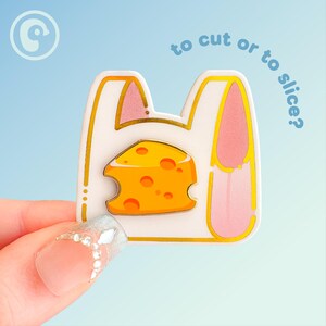 May include: A white and gold enamel pin shaped like a shopping bag with pink accents and a cheese wedge. The pin has the text "to cut or to slice?" in blue. The pin is held by a hand with a decorated fingernail.