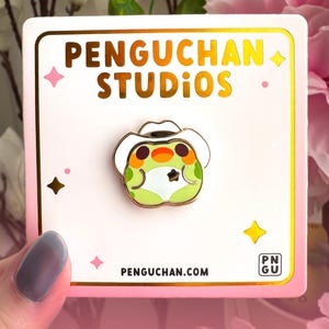 May include: A colorful enamel pin featuring a cartoon frog wearing a white cowboy hat. The pin is on a white card with gold trim, the text "PENGUCHAN STUDIOS" and "PENGUCHAN.COM". The background is pink and white.