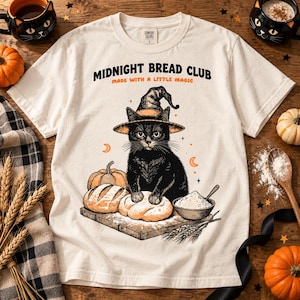 May include: A cream-colored t-shirt featuring a graphic of a black cat wearing a witch hat, kneading dough on a wooden board. The text reads MIDNIGHT BREAD CLUB MADE WITH A LITTLE MAGIC. The design includes a bowl of flour, wheat, and pumpkins.