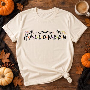 以下が含まれることがあります： クリーム色の半袖Tシャツで、黒い大文字でHALLOWEENという文字がプリントされています。文字の間には小さなカラフルなドットが配置されています。テキストの上には、ほうきに乗った2人の魔女とコウモリの黒いシルエットが描かれています。