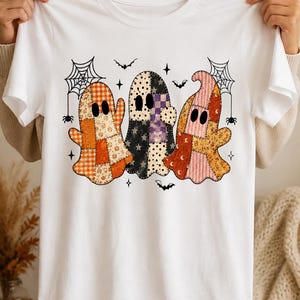 May include: A white short-sleeve t-shirt featuring a graphic of three ghosts with patchwork patterns in orange, black, purple, and pink. The design includes black spiderwebs, small spiders, bats, and star shapes surrounding the ghosts.