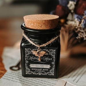 May include: A black square candle jar with a cork lid and a decorative label. The label reads "Midnight Hour" and "Midnight Muse Candles." A gold-colored charm hangs from the jar. The candle's scent is "White Peach, Charred Suede, Velvet Smoke."