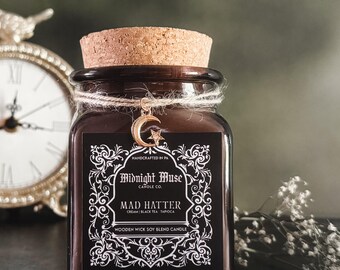 May include: A dark brown square glass jar candle with a cork lid and a gold charm. The label reads "Midnight Muse" and "Mad Hatter." The candle is tied with twine and has a wooden wick. The background includes a clock and greenery.