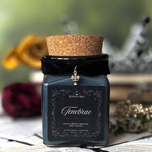 May include: A square glass candle jar with a cork lid, black velvet band, and a decorative charm. The label reads "Tenebrae" with "Black Currant, Midnight Rose, Incense" and "Coconut Soy Wax". The candle is dark blue.