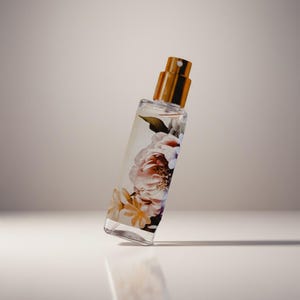 Puede incluir: Un frasco de perfume rectangular con tapa dorada y dise&ntilde;o floral. El frasco transparente est&aacute; lleno de l&iacute;quido y presenta una impresi&oacute;n de flores rosas, blancas y amarillas. El frasco est&aacute; ligeramente inclinado hacia la izquierda.
