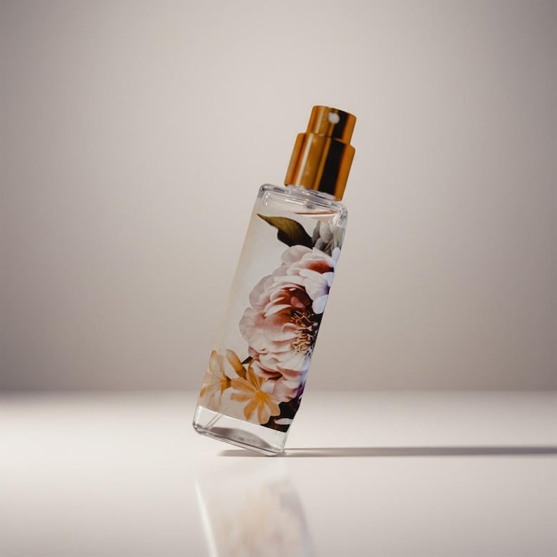 Puede incluir: Un frasco de perfume rectangular con tapa dorada y dise&ntilde;o floral. El frasco transparente est&aacute; lleno de l&iacute;quido y presenta una impresi&oacute;n de flores rosas, blancas y amarillas. El frasco est&aacute; ligeramente inclinado hacia la izquierda.