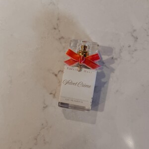Puede incluir: Un frasco de perfume rectangular con una parte superior transparente facetada y una cinta de terciopelo roja. La etiqueta dice "Velvet Cr&egrave;me" y "EAU DE PARFUM". El frasco tiene un atomizador dorado y un peque&ntilde;o dije.