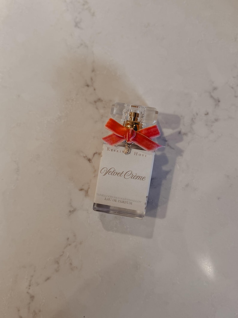 Puede incluir: Un frasco de perfume rectangular con una parte superior transparente facetada y una cinta de terciopelo roja. La etiqueta dice "Velvet Cr&egrave;me" y "EAU DE PARFUM". El frasco tiene un atomizador dorado y un peque&ntilde;o dije.