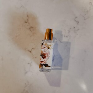 Puede incluir: Un frasco de perfume de vidrio transparente con una tapa dorada. El frasco est&aacute; decorado con un dise&ntilde;o floral con flores rosas y blancas. Contiene un l&iacute;quido transparente y est&aacute; sobre una superficie de m&aacute;rmol.