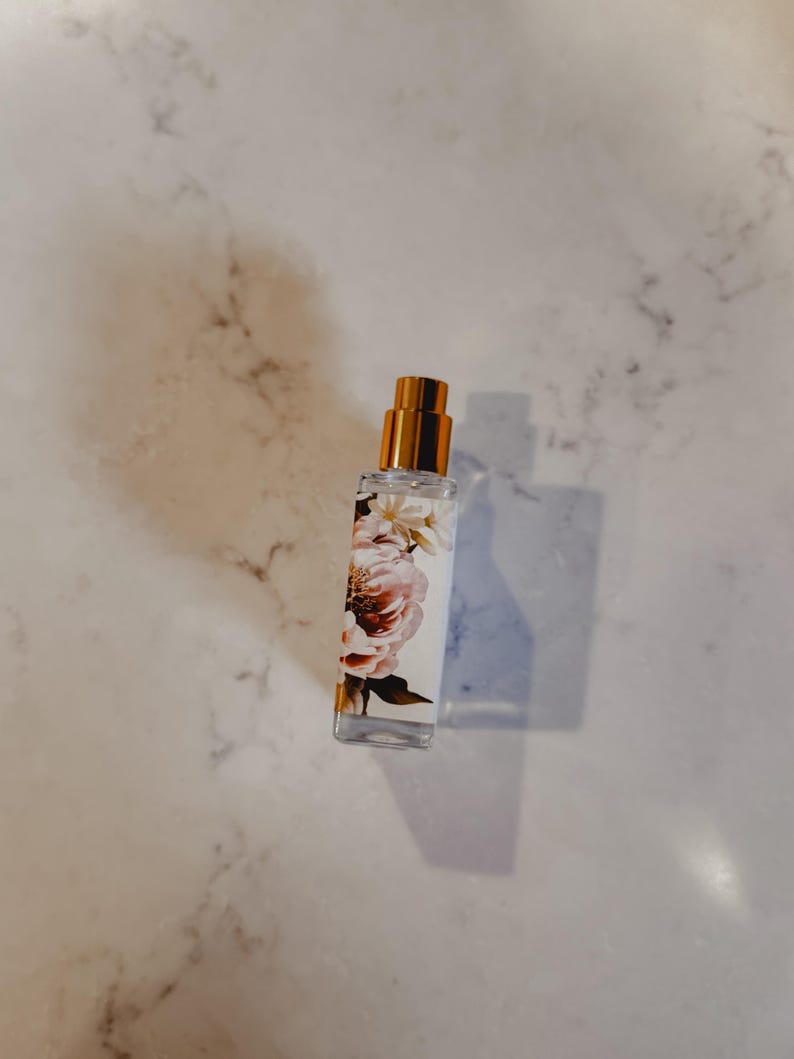 Puede incluir: Un frasco de perfume de vidrio transparente con una tapa dorada. El frasco est&aacute; decorado con un dise&ntilde;o floral con flores rosas y blancas. Contiene un l&iacute;quido transparente y est&aacute; sobre una superficie de m&aacute;rmol.