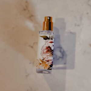 Puede incluir: Un frasco de perfume de vidrio transparente con una tapa dorada y un dise&ntilde;o floral. El frasco presenta una etiqueta blanca con flores rosas, melocot&oacute;n y amarillas. El frasco de perfume es rectangular y tiene un pulverizador.