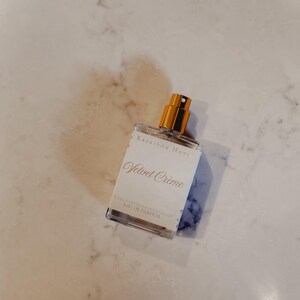Puede incluir: Un frasco de perfume rectangular transparente con tap&oacute;n dorado y etiqueta blanca. La etiqueta dice "Velvet Cr&egrave;me" en cursiva, con texto adicional debajo. El fondo es una superficie clara y veteada.