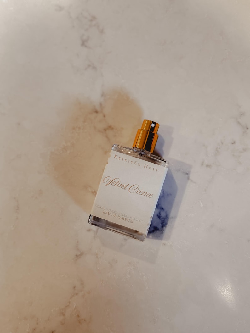 Puede incluir: Un frasco de perfume rectangular transparente con tap&oacute;n dorado y etiqueta blanca. La etiqueta dice "Velvet Cr&egrave;me" en cursiva, con texto adicional debajo. El fondo es una superficie clara y veteada.